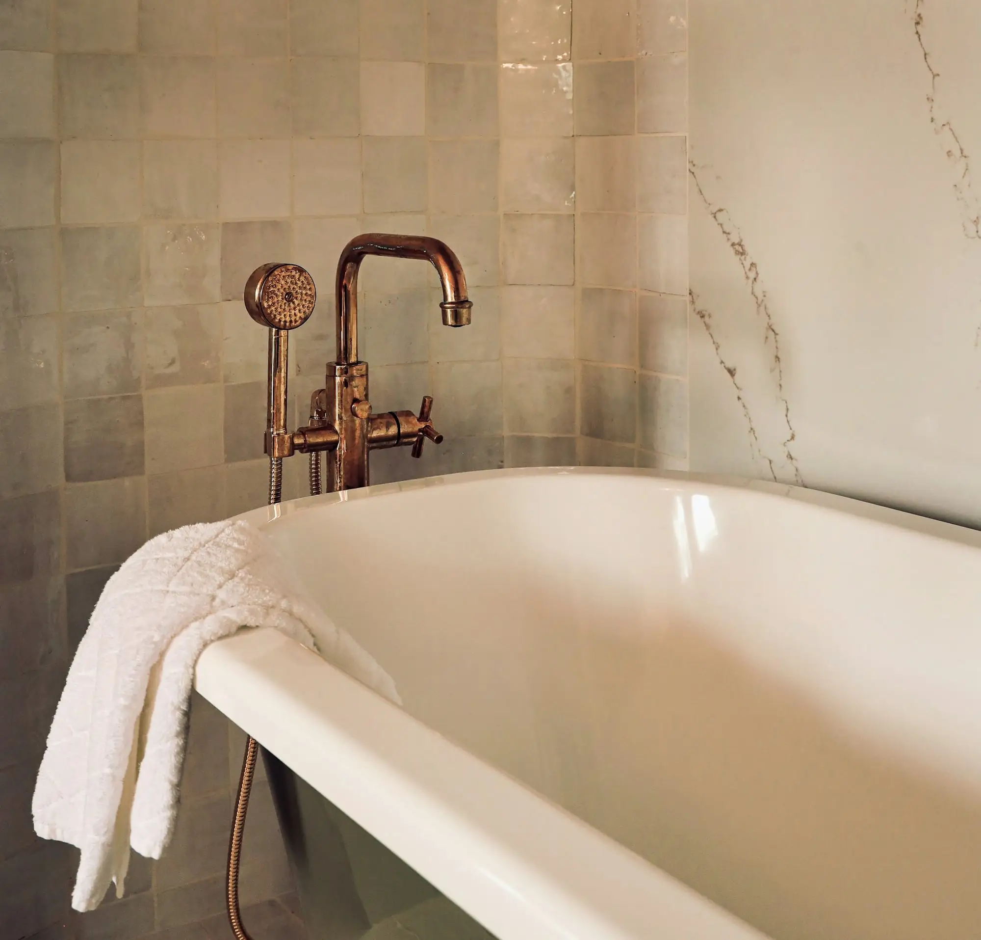Copper-finish tub filler and handheld shower beside a white bathtub with veined stone tile behind