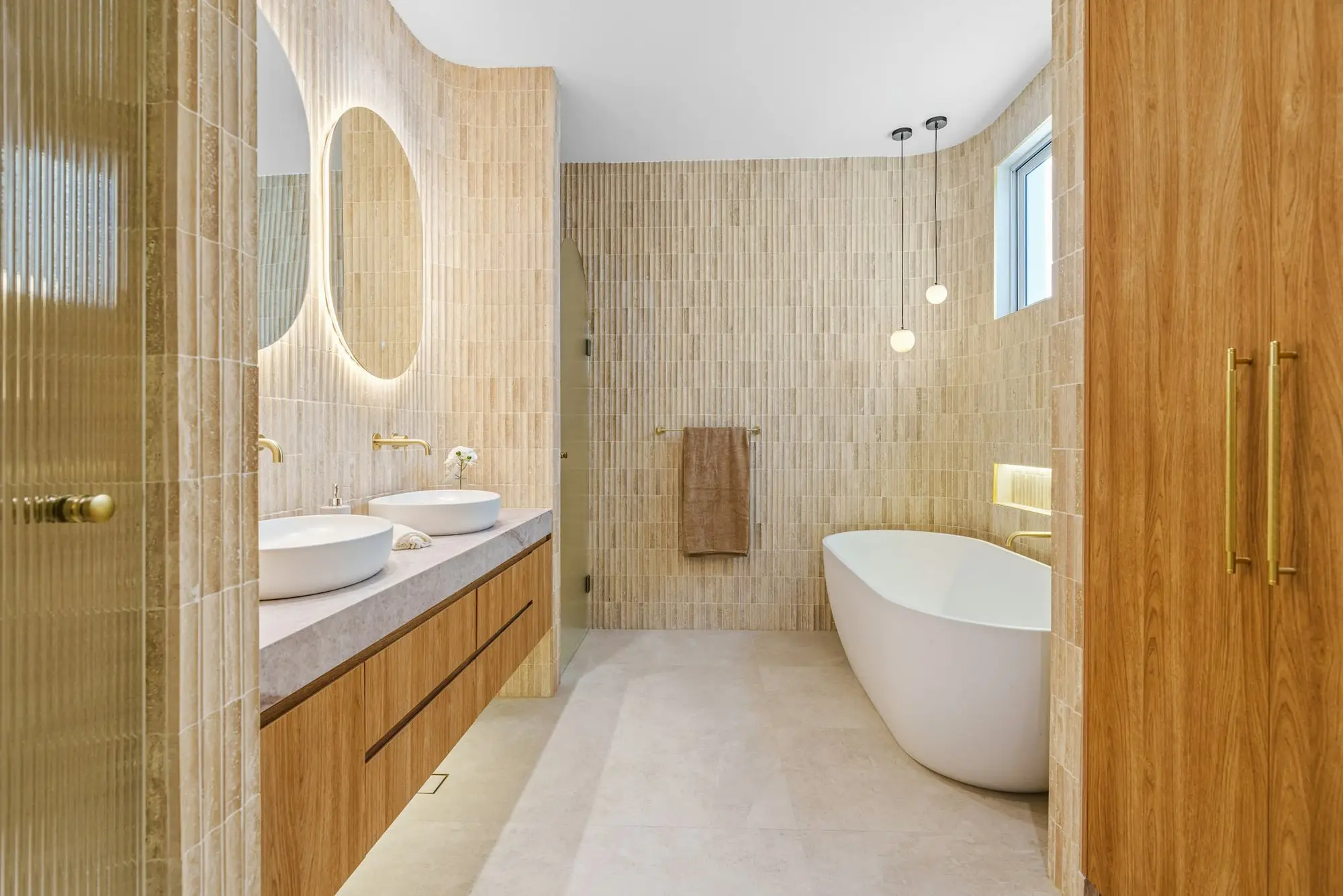 Oak double vanity, ribbed glass partition and a freestanding tub beneath a round backlit mirror