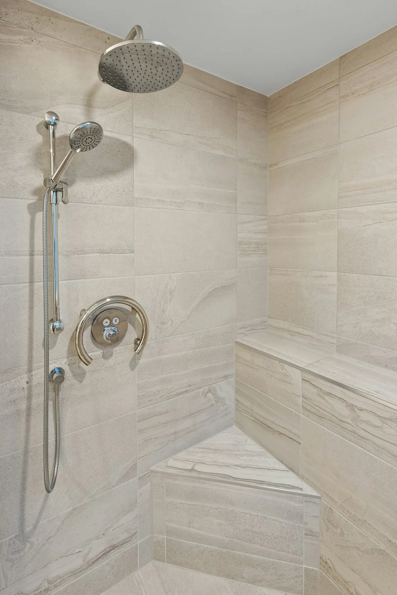 Tiled shower in warm stone-look porcelain with a built-in corner bench, rain head and handheld shower