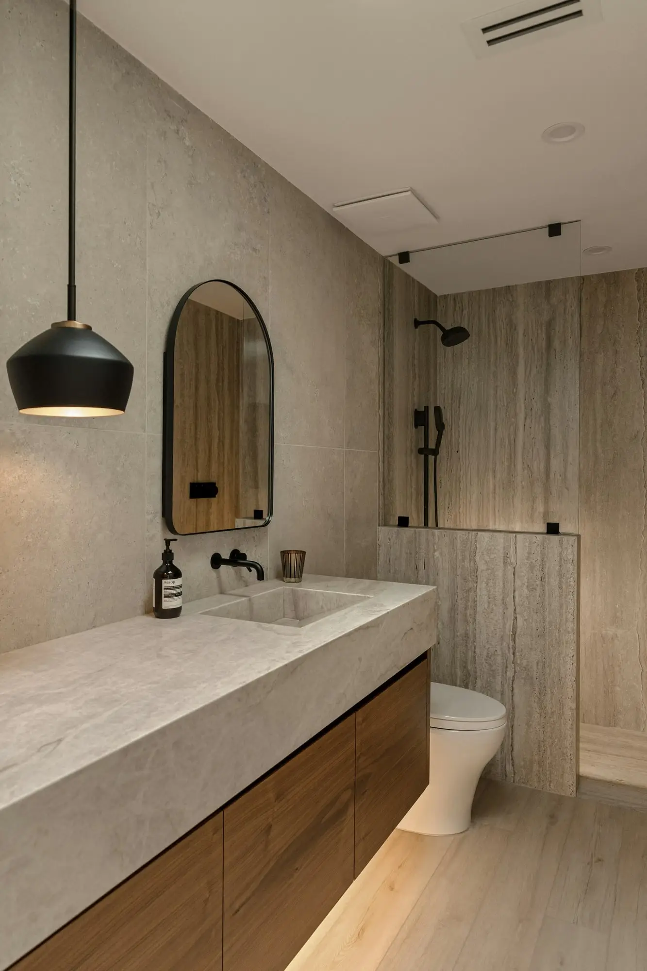 Travertine-look vanity with a wood base, arched mirror and black pendant light in a compact bathroom